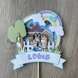 K&ouml;nnte beinhalten: Ein blauer, gr&uuml;ner und lilafarbener Kuchentopper mit den Figuren aus der Show Bluey. Der Topper hat ein Banner mit der Aufschrift "LOUIS" und einen Regenbogen mit der Zahl 3.