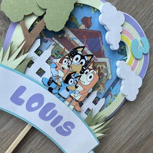 K&ouml;nnte beinhalten: Ein lila und blauer Tortendekor mit einem Regenbogen, Wolken und einem Haus mit einem Fenster. Das Haus enth&auml;lt drei Cartoon-Hunde. Der Topper hat ein Banner mit der Aufschrift "Louis" in lila Buchstaben.
