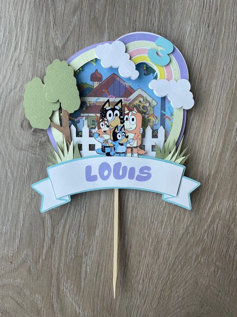K&ouml;nnte beinhalten: Ein blauer, gr&uuml;ner und lilafarbener Kuchentopper mit den Figuren aus der Show Bluey. Der Topper hat ein Banner mit der Aufschrift "LOUIS" und einen Regenbogen mit der Zahl 3.