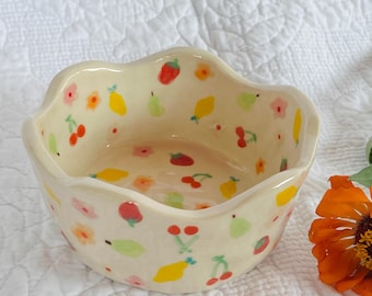 Op de afbeelding: Een kleine, crèmekleurige keramische kom met een geschulpte rand. De kom is versierd met kleurrijke illustraties van fruit en bloemen. Een paar oranje en roze bloemen liggen naast de kom op een wit, gewatteerd oppervlak.