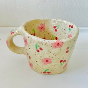 May include: A handmade ceramic mug with a cream-colored speckled finish. The mug is adorned with pink floral and red cherry designs. It has a rounded handle and a unique, irregular shape, perfect for enjoying your favorite beverage.