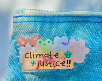 May include: A blue denim pencil case with a clear sticker featuring cartoon bears, a frog, and the words "climate justice!!" in a colorful design. The sticker includes a rainbow and a globe.