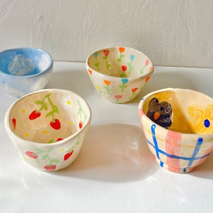 May include: Four small, hand-painted ceramic bowls. One bowl has a blue cloud design, another has a strawberry pattern, and a third has a floral design. The fourth bowl has a butterfly and flower design with a pink and blue plaid pattern.