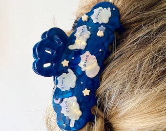 May include: A deep blue translucent hair claw clip featuring an iridescent finish. The clip is decorated with small, white, cat-shaped figures, pale yellow stars, and a tiny purple crescent moon, all arranged across the surface of the accessory.