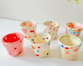 May include: A collection of seven small, hand-painted ceramic cups. Each cup features a unique design, including cherries, hearts, strawberries, and floral patterns. The cups are in various colors, including pink, red, and white.