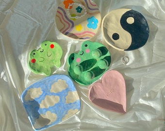 May include: A collection of handmade ceramic trinket dishes in various shapes and designs. Includes a round dish with a yin-yang symbol, frog-shaped dishes, a heart-shaped dish with "XOXO", and a cloud-patterned dish. The dishes are painted in bright colors.