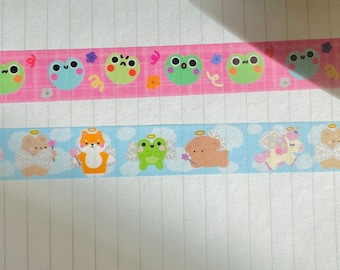 May include: Two rolls of decorative washi tape. One roll is pink with green frog illustrations. The other roll is blue with illustrations of a fox, a frog, a sheep, and a unicorn. The tape is on a white lined paper.
