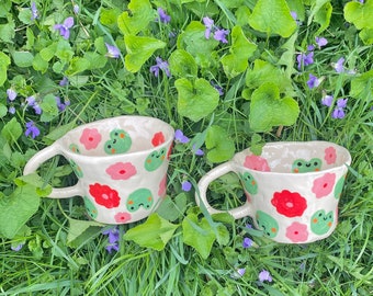May include: Two ceramic mugs with white backgrounds, adorned with red and pink flower and green frog designs. The mugs have curved handles and are set on a bed of green grass and purple flowers.