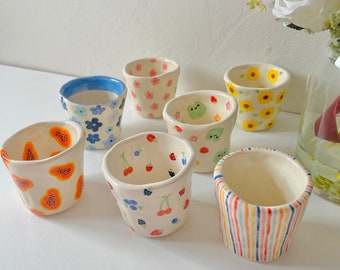 May include: A collection of seven small, hand-painted ceramic cups. Each cup features a unique design, including floral patterns, fruit illustrations, and colorful stripes. The cups are white with various colors and designs.
