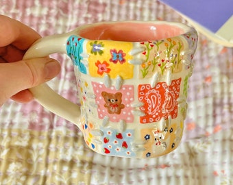 May include: A ceramic mug with a patchwork design featuring various patterns and colors, including flowers, strawberries, and a teddy bear. The interior is pink, and the handle is off-white.
