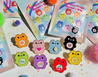 May include: An assortment of colorful enamel pins and mystery bags. The pins are frog-shaped with various designs, including a heart, moon, and rainbow. The bags are labeled "Rainbow Frogs Mystery Bag" with a cloud and rainbow design.