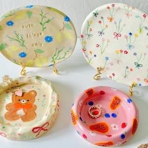 May include: Four decorative ceramic trays and dishes. One is yellow with blue flowers and text, another has a floral pattern, a third has a bear design, and the last is pink with fruit. Jewellery is displayed on two of the trays.