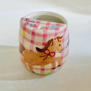 May include: A ceramic cup featuring a pink and white checkered pattern with a hand-painted brown horse illustration. The rim includes the text Ride easy in black script, a small circular hole, and a small blue flower accent on the side.