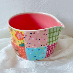 Pode incluir: Uma tigela de cerâmica com interior rosa brilhante e bico para despejar. O exterior apresenta um design de patchwork colorido com vários padrões, incluindo flores vermelhas, morangos pequenos sobre rosa, xadrez verde, um urso castanho e flores azuis.