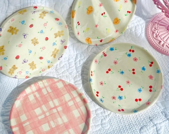 May include: Four decorative ceramic plates with various designs. One plate features a pink and white plaid pattern. The other plates have cream-coloured backgrounds with illustrations of bears, cherries, and flowers. A pink vanity mirror is in the background.