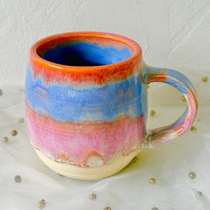 May include: A handmade ceramic mug with a colorful, layered design. The mug features bands of blue, orange, and pink hues, with a cream-colored base and a matching handle. The mug is designed for hot beverages.