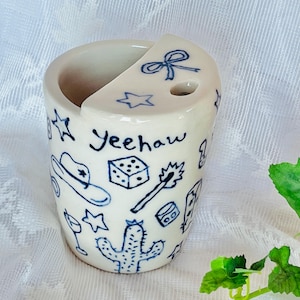 Peut inclure: Une tasse en céramique blanche avec un dessus incurvé et une découpe. La tasse est décorée de dessins bleus d'un chapeau de cowboy, d'étoiles, d'un cactus et du mot "Yeehaw". La tasse a un petit trou sur le dessus.