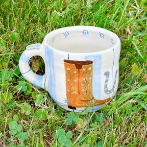 May include: A ceramic mug with a white base, featuring light blue vertical stripes and a painted orange cowboy boot with small white flowers. The mug includes a grey horseshoe graphic, a small grey heart on the handle, and tiny blue flowers painted along the inner rim.