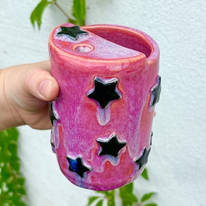 May include: A cylindrical ceramic mug featuring a vibrant pink and purple mottled glaze. The exterior is decorated with multiple raised, glossy black star-shaped appliqués. The top rim has a distinct slanted cut-out design with a small circular hole.