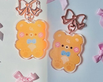 May include: Two yellow bear-shaped keychains with rose gold bow-shaped clasps and blue bow ties. The keychains are surrounded by small pink satin bows on a white and light blue background. The keychains have a cute, cartoonish design.