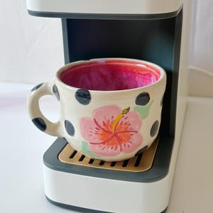 May include: A ceramic mug with a cream-colored exterior featuring black polka dots and a large pink hibiscus flower design. The interior of the mug is glazed in a vibrant, deep magenta color. The mug has a curved handle and sits on a coffee machine.
