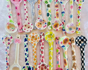May include: A collection of decorative ceramic spoons with various colorful patterns. Designs include strawberries, hearts, and floral motifs. The spoons are hand-painted with a glossy finish, showcasing a range of pinks, greens, blues, and yellows.