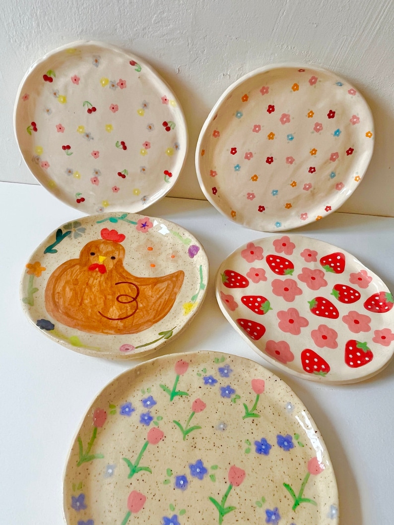 Op de afbeelding: Een verzameling van vijf handbeschilderde keramische borden. De borden hebben verschillende ontwerpen, waaronder bloempatronen, kersen, aardbeien en een kip. Het kleurenpalet omvat roze, rood, geel en blauw op een gespikkelde beige achtergrond. De borden hebben onregelmatige, organische vormen.