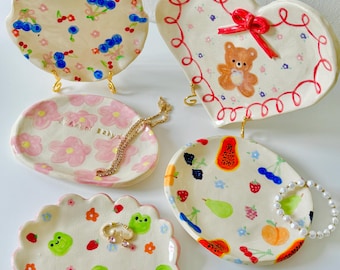 May include: A collection of hand-painted ceramic trinket dishes. The dishes feature various designs, including cherries, a teddy bear, flowers, frogs, and fruit. Each dish is a unique shape and rests on a gold-colored stand. A necklace and bracelets are displayed on the dishes.