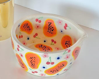 May include: A white ceramic bowl with a spout, adorned with hand-painted papaya and cherry designs. The papaya is orange with black seeds, and the cherries are red. Pink flowers and blue dots complete the design.