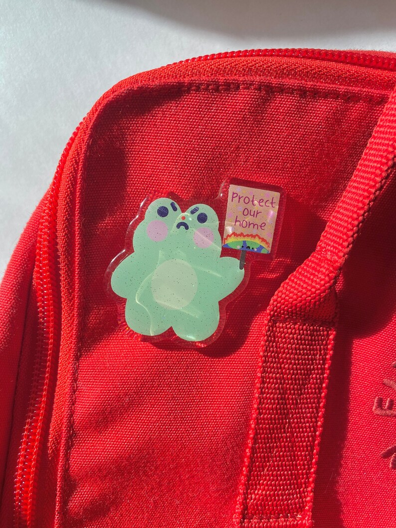 May include: A light green frog-shaped pin with glitter accents, holding a sign that reads "Protect our home" with a rainbow. The pin is attached to a red bag.