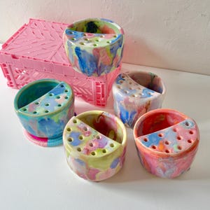 May include: Four round ceramic brush holders with a marbled, multi-colored glaze in shades of blue, pink, yellow, and green. Each holder features a flat, semi-circular top section with multiple small circular holes for organizing brushes.