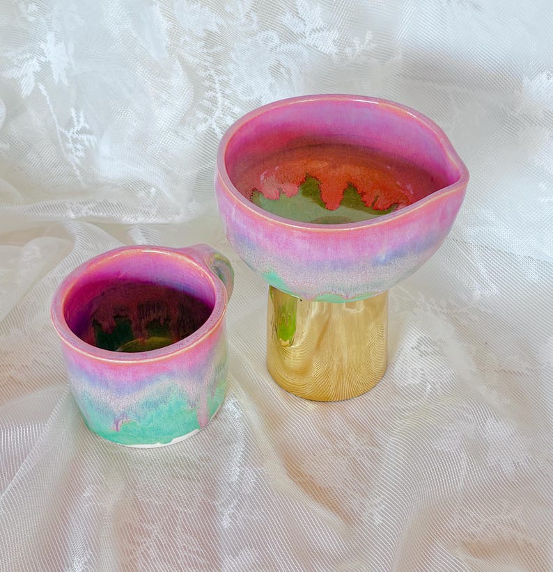 Pu&ograve; includere: Due pezzi in ceramica con smalto iridescente. Uno &egrave; una piccola tazza con manico, l'altro &egrave; un pezzo a forma di ciotola su una base dorata. Lo smalto passa dal rosa al verde e all'oro.