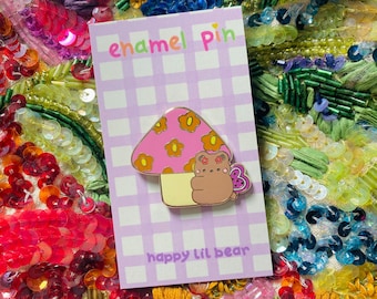 May include: An enamel pin featuring a mushroom design with a brown bear and butterfly. The pin is on a card with the text "enamel pin" and "happy lil bear" against a purple and white checkered background. The mushroom has a pink cap with orange flowers.