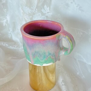Pu&ograve; includere: Una tazza in ceramica fatta a mano con una smaltatura colorata unica. L'interno della tazza &egrave; rosa, che sfuma in tonalit&agrave; verdi e blu all'esterno. La tazza &egrave; alta circa 10 cm e ha una base dorata.