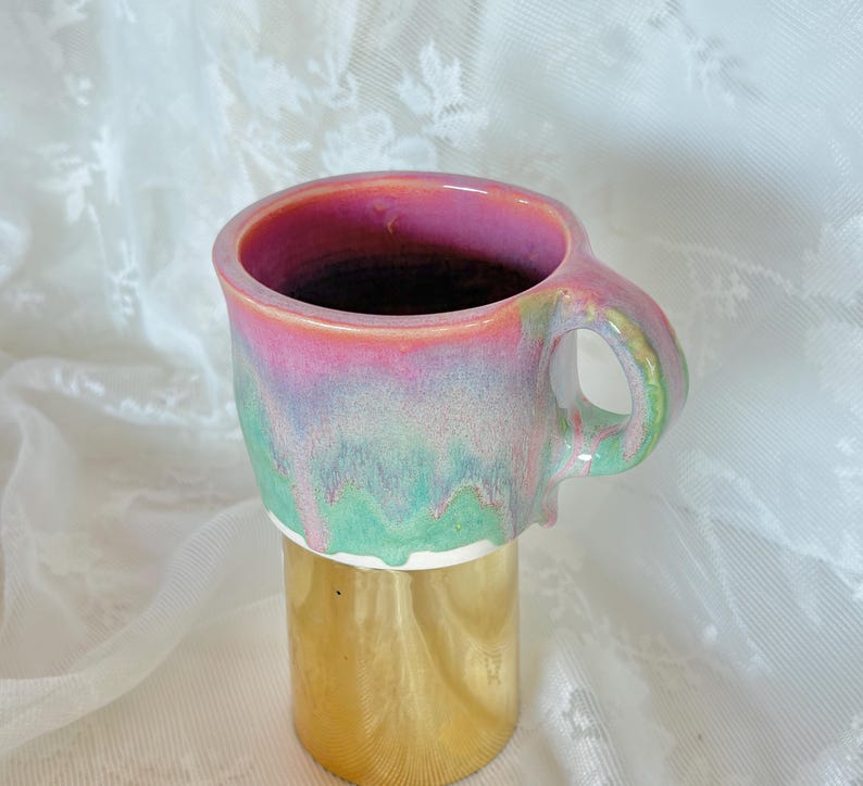 Pu&ograve; includere: Una tazza in ceramica fatta a mano con una smaltatura colorata unica. L'interno della tazza &egrave; rosa, che sfuma in tonalit&agrave; verdi e blu all'esterno. La tazza &egrave; alta circa 10 cm e ha una base dorata.