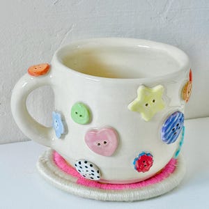 May include: A white ceramic mug decorated with various colorful, raised button-shaped embellishments including a yellow star, a pink heart, and blue, green, and polka-dot circles. The mug rests on a round, cream-colored coaster with a bright pink center.