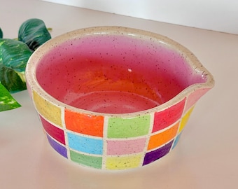 May include: A colorful ceramic mixing bowl with a spout. The bowl features a checkerboard pattern of various colors, including red, orange, yellow, green, and blue. The interior is a gradient of pink and orange.