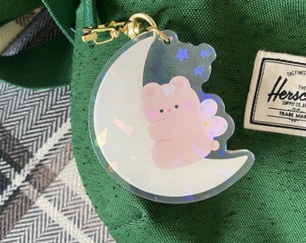 May include: A clear acrylic keychain featuring a pink bear sitting on a crescent moon with iridescent accents. The keychain is attached to a green bag with a gold-colored clasp. A Herschel Supply Co. tag is visible.