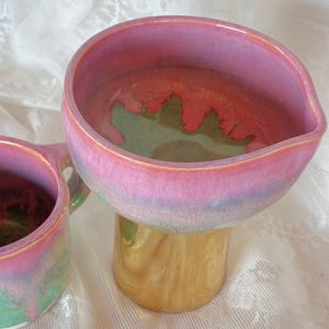 Pu&ograve; includere: Una ciotola in ceramica fatta a mano con una base a piedistallo color oro. La ciotola presenta una vibrante tavolozza di colori con tonalit&agrave; rosa, verde e blu. L'interno della ciotola ha un design unico e organico.