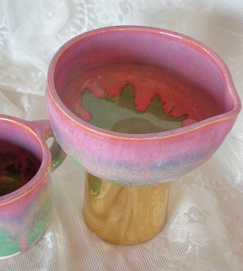 Pu&ograve; includere: Una ciotola in ceramica fatta a mano con una base a piedistallo color oro. La ciotola presenta una vibrante tavolozza di colori con tonalit&agrave; rosa, verde e blu. L'interno della ciotola ha un design unico e organico.