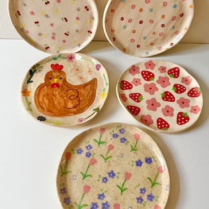 Op de afbeelding: Een verzameling van vijf handbeschilderde keramische borden. De borden hebben verschillende ontwerpen, waaronder bloemmotieven, aardbeien en een kip. Het kleurenpalet omvat roze, rood, geel en blauw op een gespikkelde beige achtergrond.