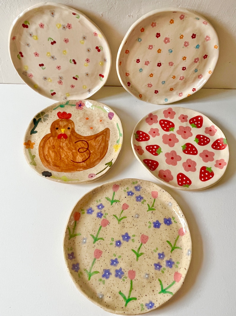 Op de afbeelding: Een verzameling van vijf handbeschilderde keramische borden. De borden hebben verschillende ontwerpen, waaronder bloemmotieven, aardbeien en een kip. Het kleurenpalet omvat roze, rood, geel en blauw op een gespikkelde beige achtergrond.