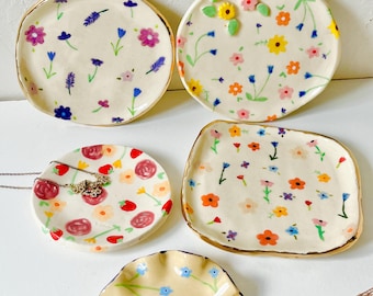 May include: A collection of five hand-painted ceramic dishes with floral designs. The dishes vary in shape, including round and rectangular, and feature colorful flowers, gold rims, and delicate details. Ideal for jewelry or small items.