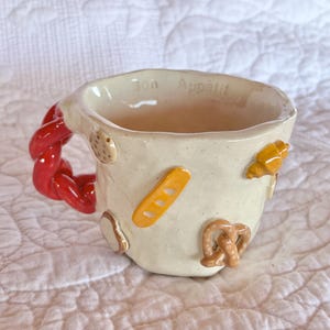 May include: Cream-colored ceramic mug with a red, braided handle. The mug is decorated with miniature bread-themed embellishments, including a pretzel, croissant, and baguette. The words "Bon Appétit" are written on the rim.