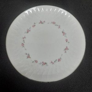 May include: A white ceramic plate with a scalloped edge and a delicate floral design in pink and green. The plate has a subtle embossed pattern.
