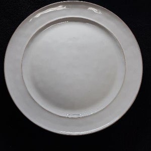 May include: A white ceramic dinner plate with a slightly raised rim. The plate has a subtle, off-white glaze.