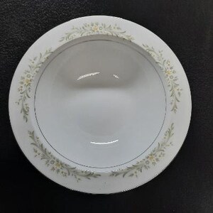 May include: A white ceramic bowl with a green and white floral design around the rim. The bowl is empty and has a slight sheen.
