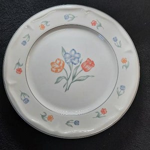 May include: A white ceramic dinner plate with a floral design. The plate features a cluster of orange, blue, and pink flowers with green leaves. The rim of the plate is decorated with a repeating pattern of small blue and orange flowers.