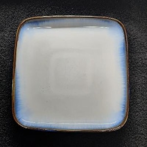 May include: A square ceramic plate with a white glaze and a blue rim. The plate has a subtle brown glaze around the edge.