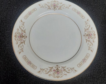 Può includere: Un piatto da portata in ceramica bianca con un design dorato e floreale. Il piatto ha un bordo dorato e un motivo floreale con fiori rosa e marroni.
